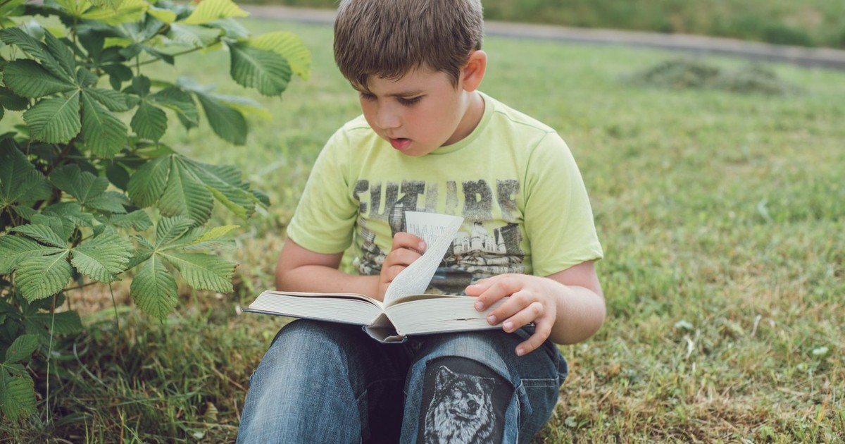 A young boy sits on grass outdoors, engrossed in reading a book.