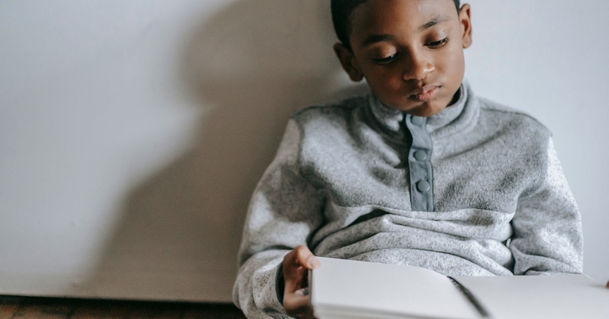 Concentrated African American boy looking at opened copybook while sitting on parquet at home