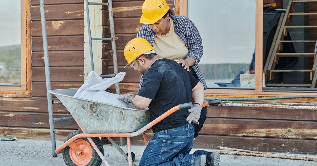 A construction worker receiving help while kneeling with a wheelbarrow on a job site.