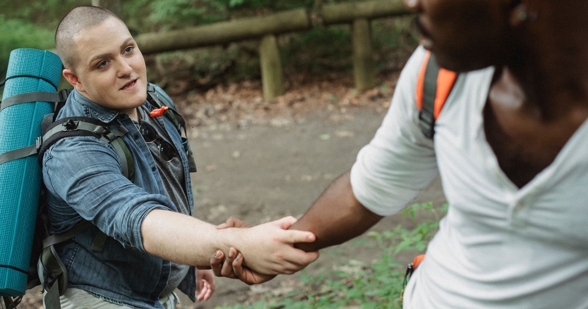 Unrecognizable black adventurer providing hand for happy plump male friend with big backpack and rolled rug in woodland in daytime