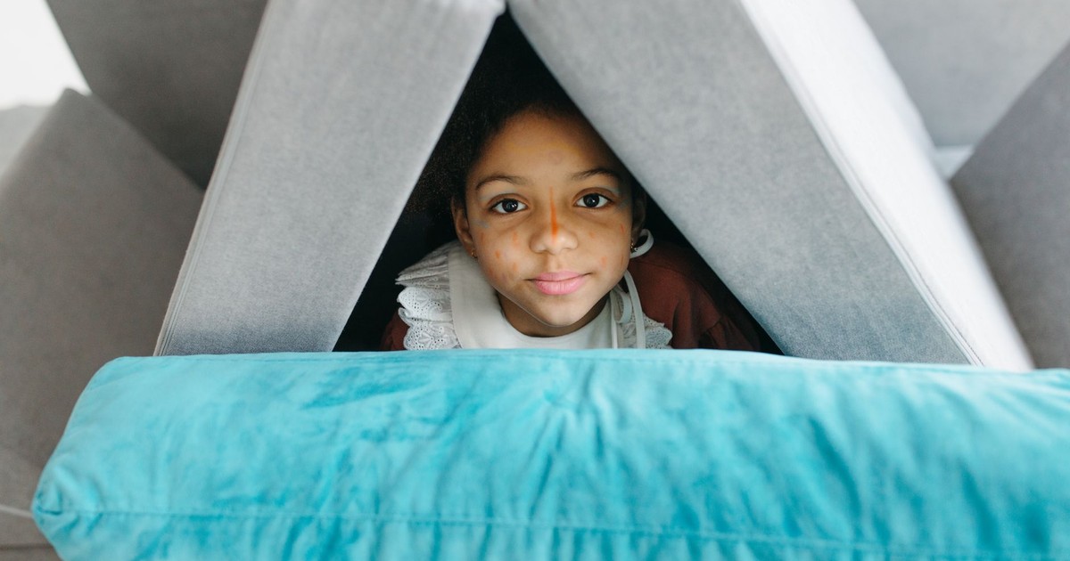 A young girl with face paint hides inside a cozy pillow fort, peeking out with curiosity.