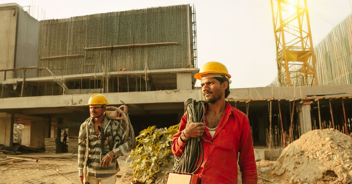 Two construction workers wearing helmets at a site in India with a crane and building.