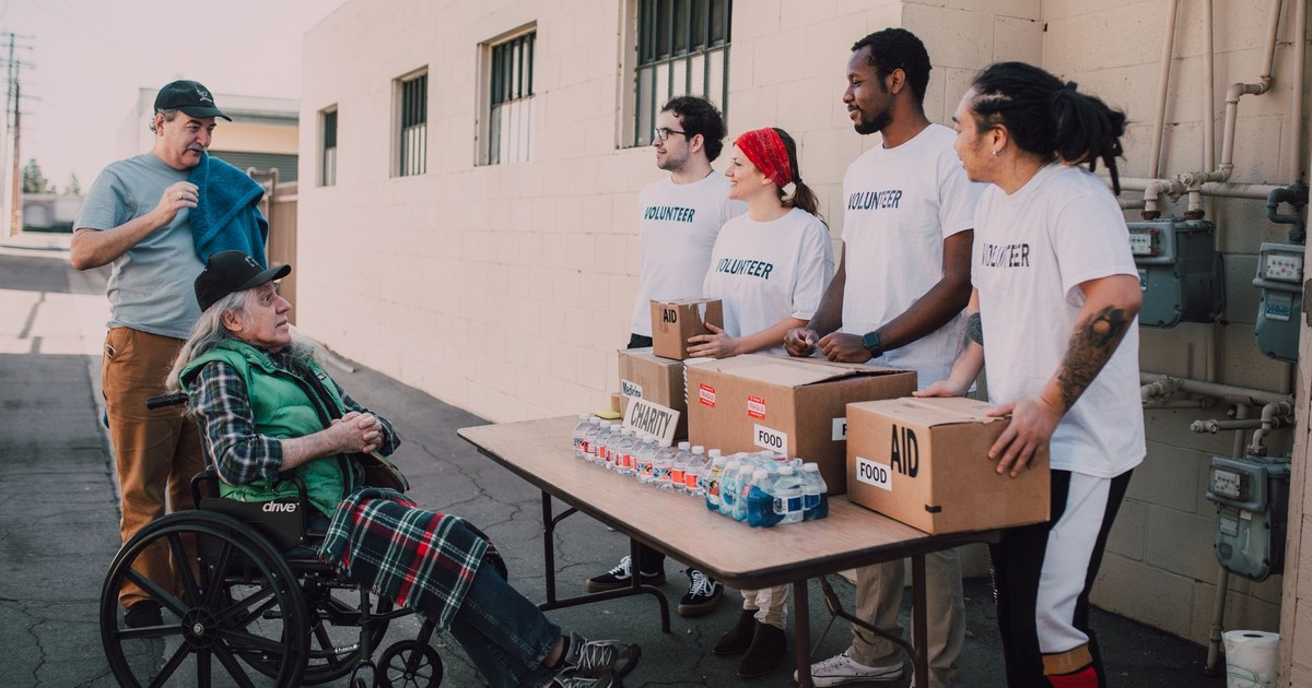 Volunteers assist a senior adult in a wheelchair by distributing aid packages outdoors.