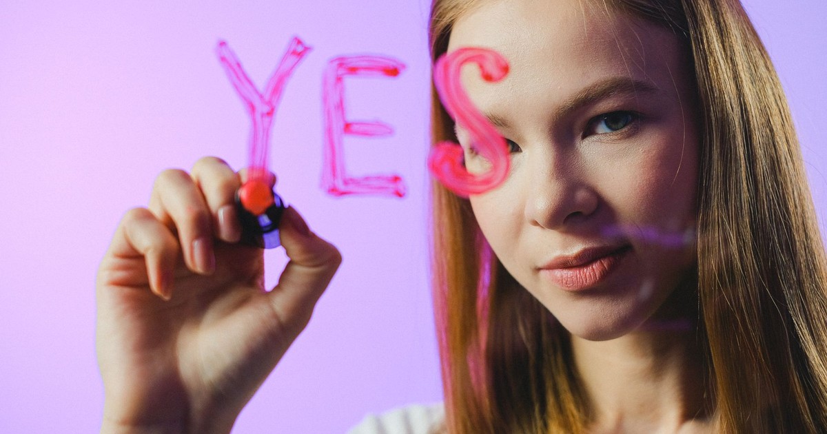 Teenage girl writing the word 'Yes' on glass with marker in studio setting.