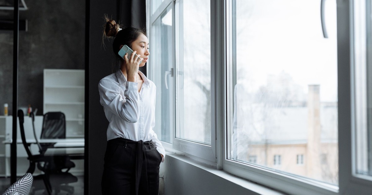 Asian woman in office clothes making a phone call while gazing outside a large window.