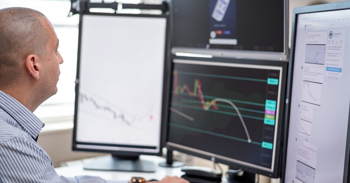 Stock trader analyzing financial graphs on multiple computer monitors in an office setting.