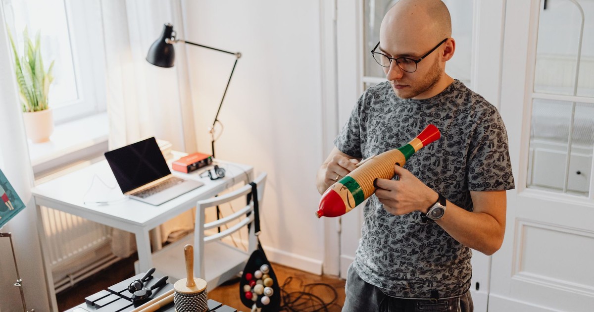 A man in casual attire plays a wooden guiro in a modern home studio setting.