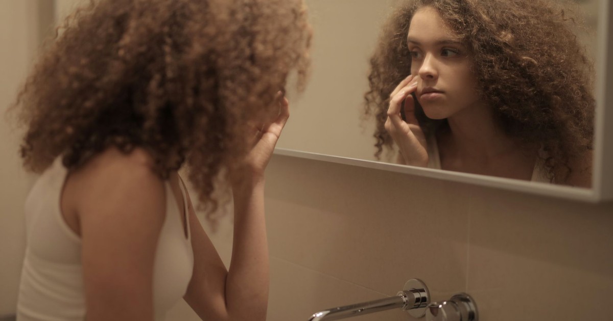 Teenage girl with curly hair examines her face in the bathroom mirror, reflecting contemplation.