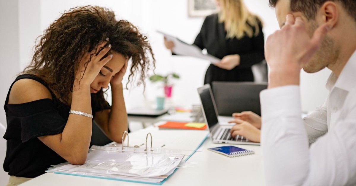African American woman stressed at office desk with colleagues nearby, conveying workplace pressure.