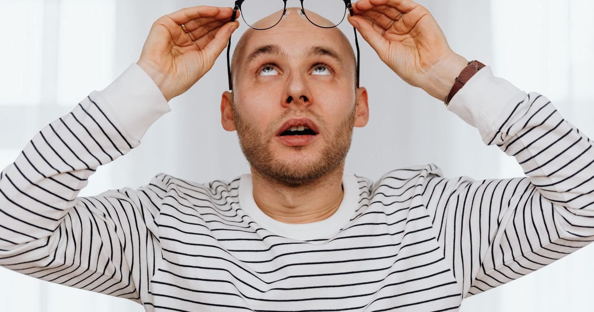 Young bearded man in striped sweater adjusting eyeglasses indoors with a surprised expression.