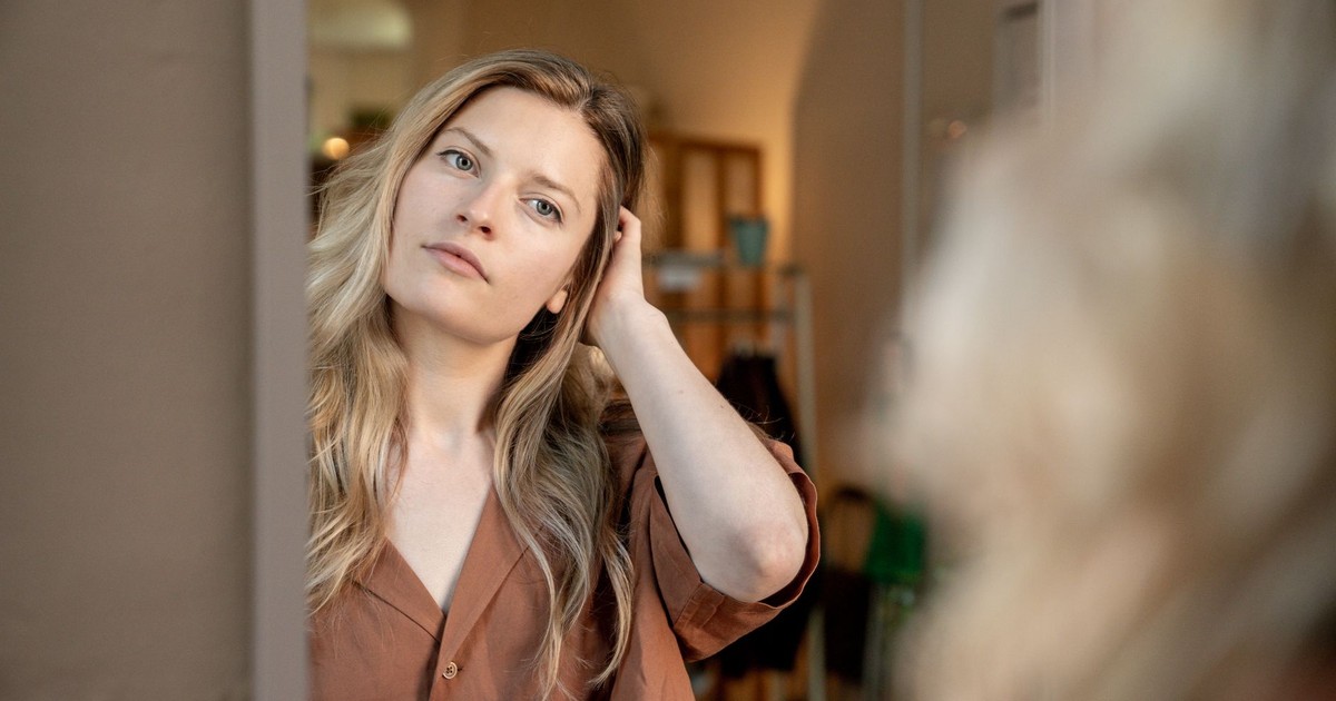 A thoughtful woman holding a coffee cup looks at her reflection in the mirror in a cozy indoor setting.