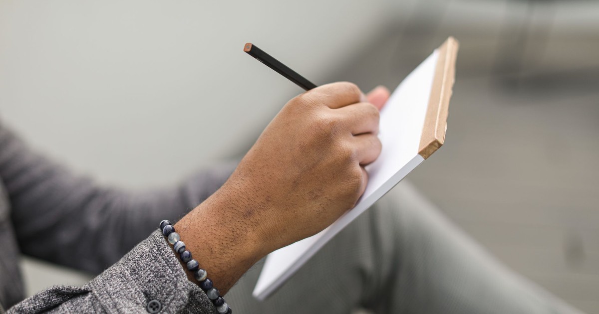 An adult hand writes in a notebook with a pencil, wearing a beaded bracelet and gray plaid sleeves.
