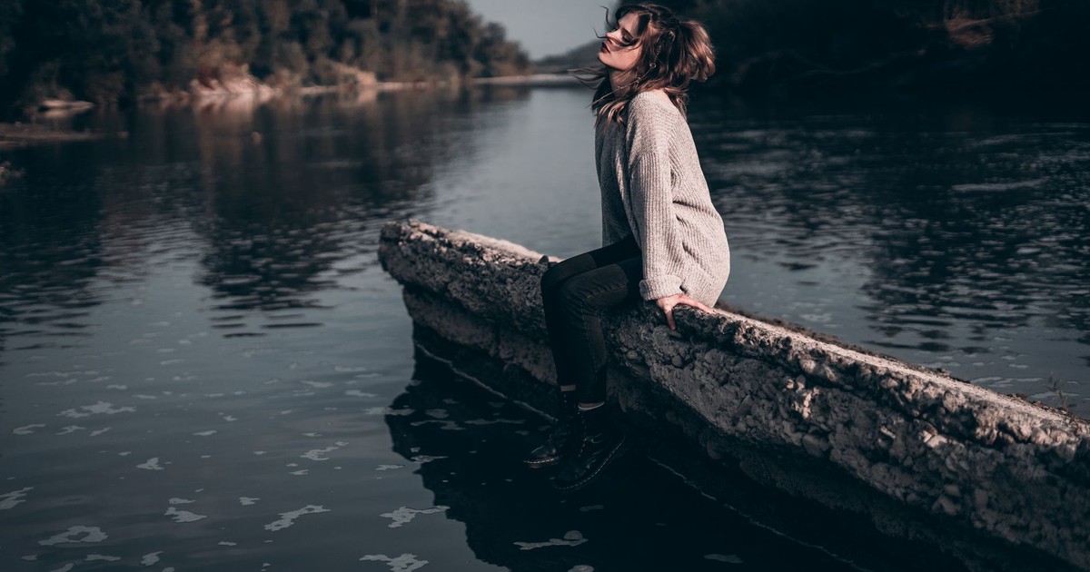 A woman enjoys a peaceful moment sitting on a riverside barrier during sunset.