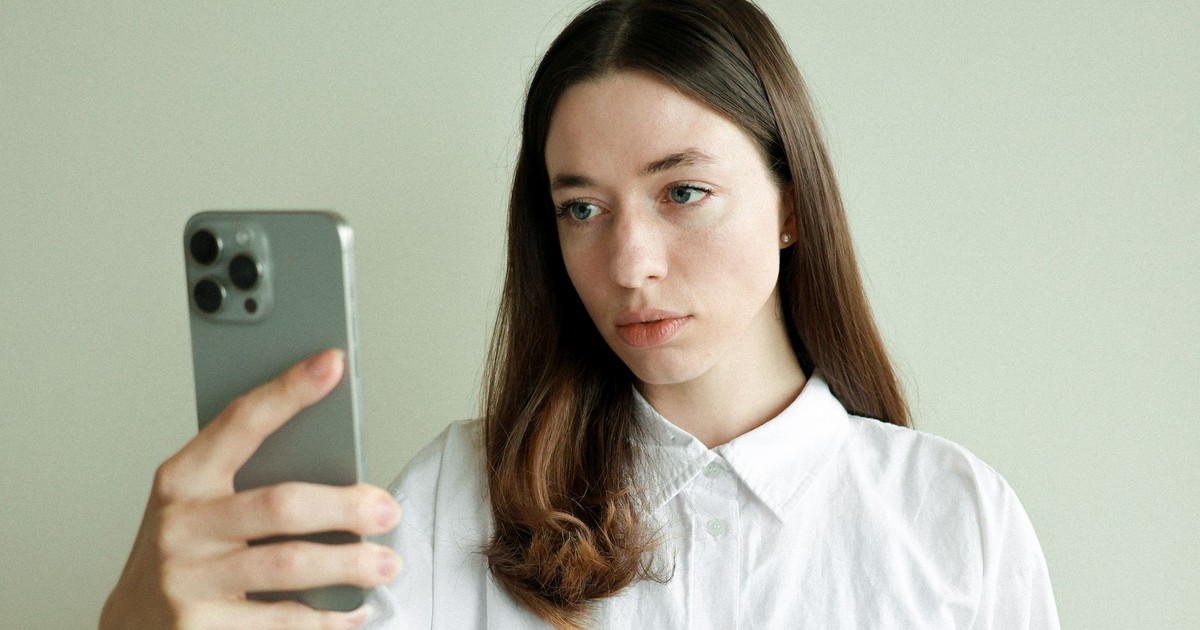 Young woman with long hair takes selfie using smartphone indoors, neutral background.
