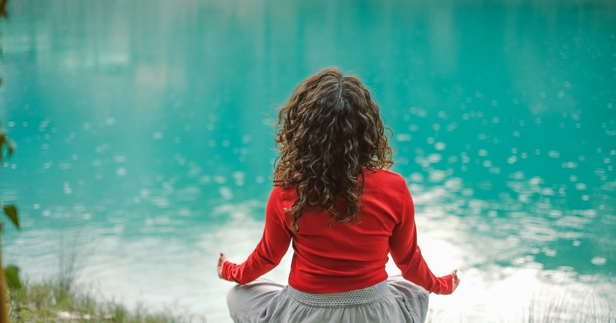 A woman in red meditates by a tranquil blue lake, reflecting calm and serenity.