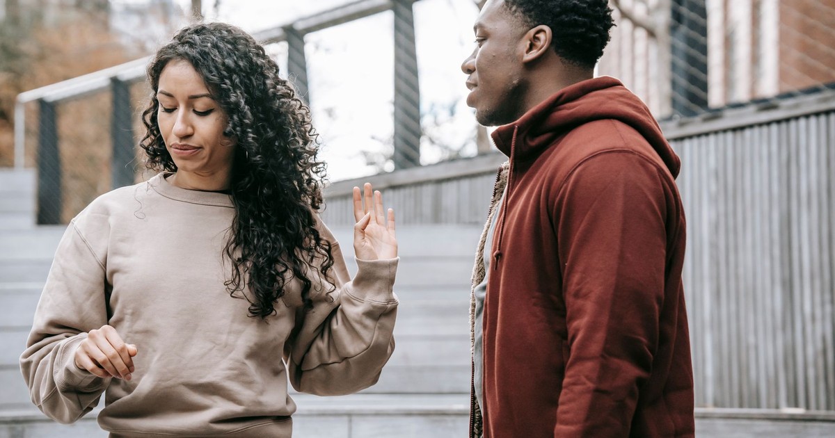 Upset ethnic girlfriend with raised hand and concerned African American boyfriend breaking up on street against metal grid on background
