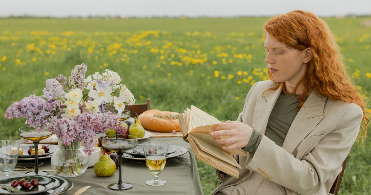 Woman with red hair reading at a beautifully set outdoor picnic in a summer field.