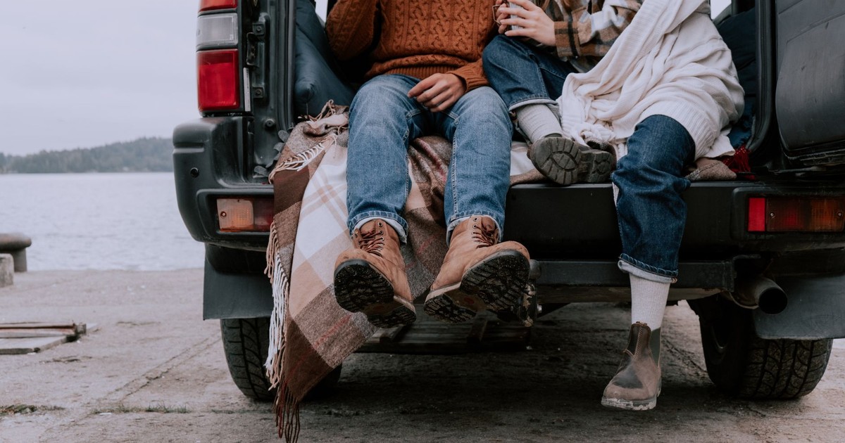 A couple enjoys a warm drink while sitting in an SUV trunk by the serene lakeside on a chilly day.