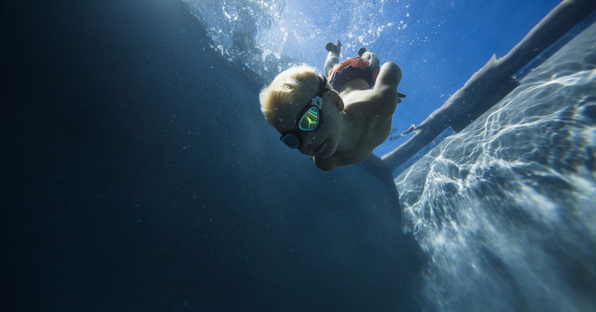 A young boy wearing goggles dives underwater in a sunny pool, embodying summer fun.