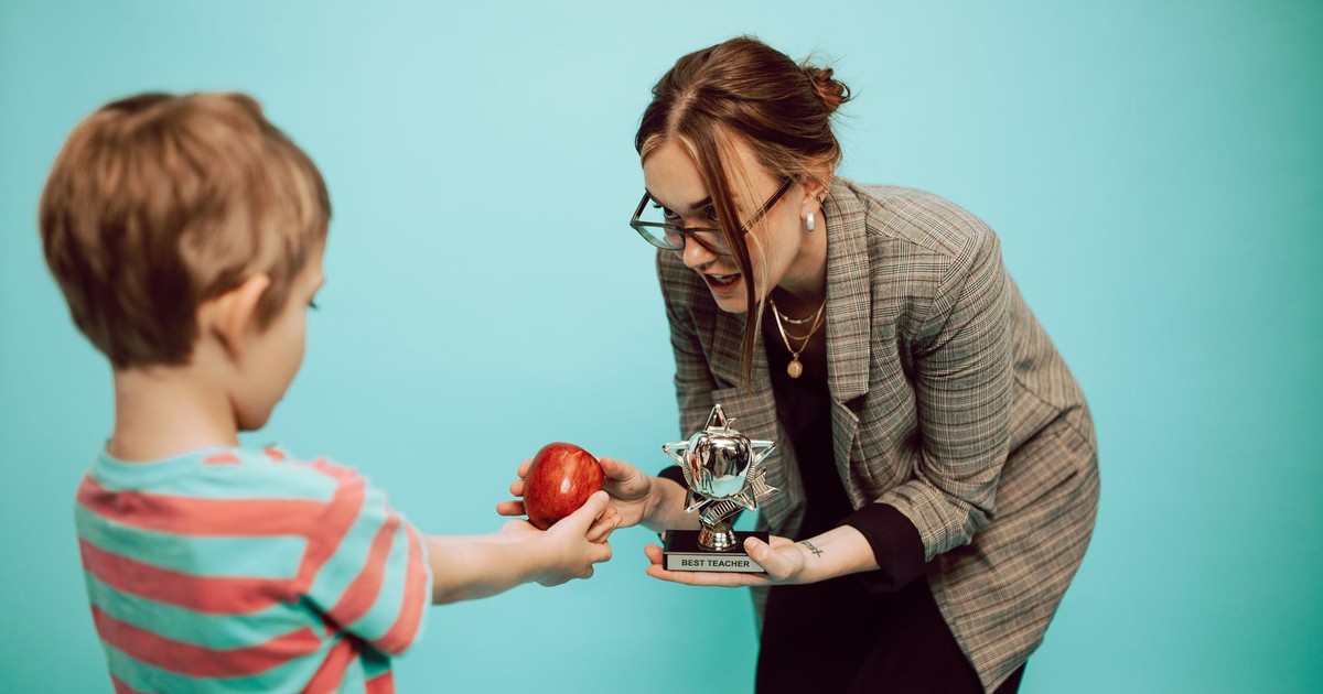 A child offers an apple to a teacher holding a "Best Teacher" trophy in a classroom scene.
