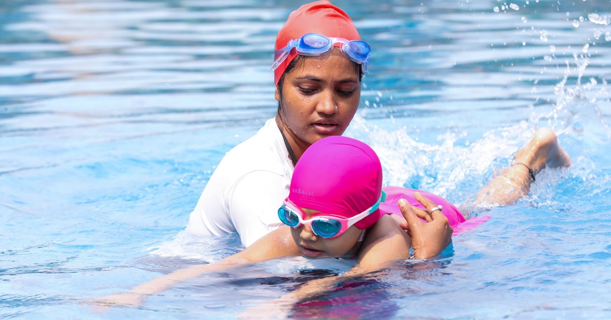 Instructor guiding a child in swimming pool, wearing swim caps and goggles.