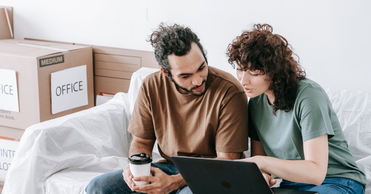 Young couple sits on a sofa using a laptop during a move, surrounded by boxes and sipping coffee.