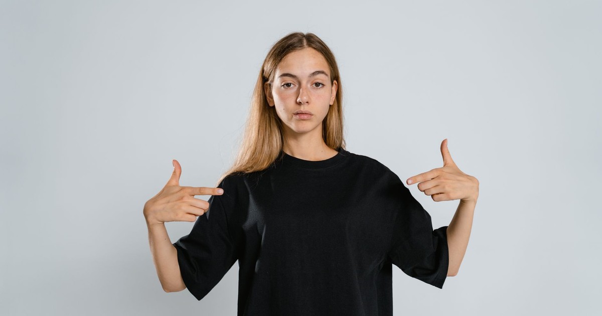 Portrait of a woman in a black t-shirt pointing at herself on a white background.