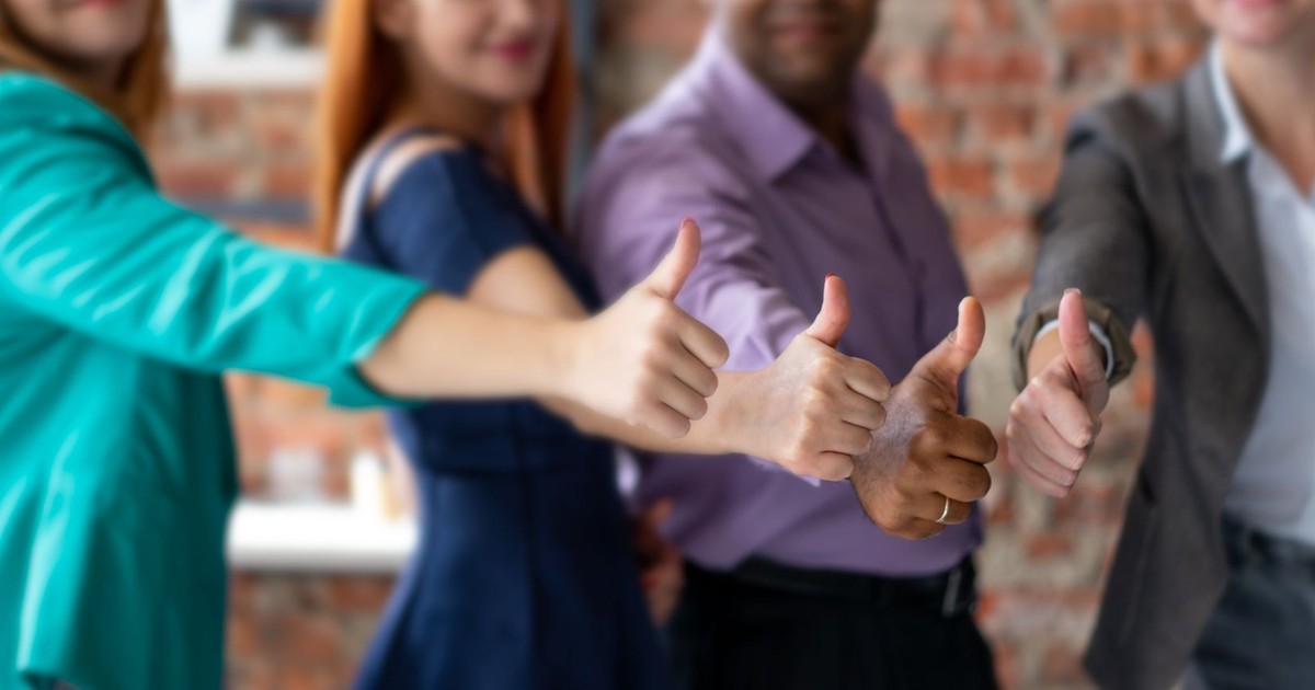 Unrecognizable cheerful colleagues in formal clothes showing thumbs up gesture while standing in modern office during teamwork on blurred background
