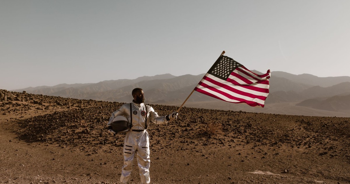 An astronaut in a space suit stands in a desert landscape holding an American flag.