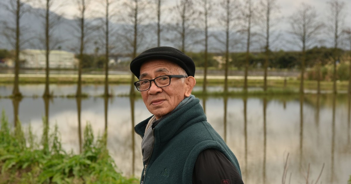 Senior man in wool vest by a serene lake in Taiwan during daytime.