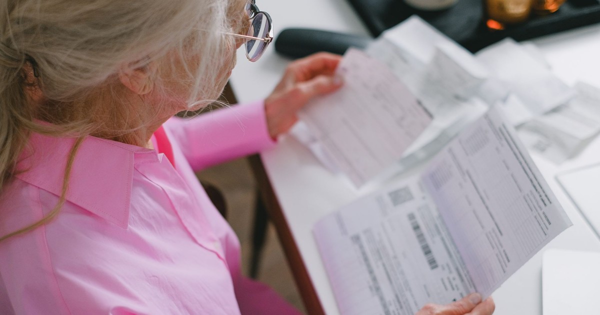 An elderly woman in glasses holds and reads important papers at a table indoors.