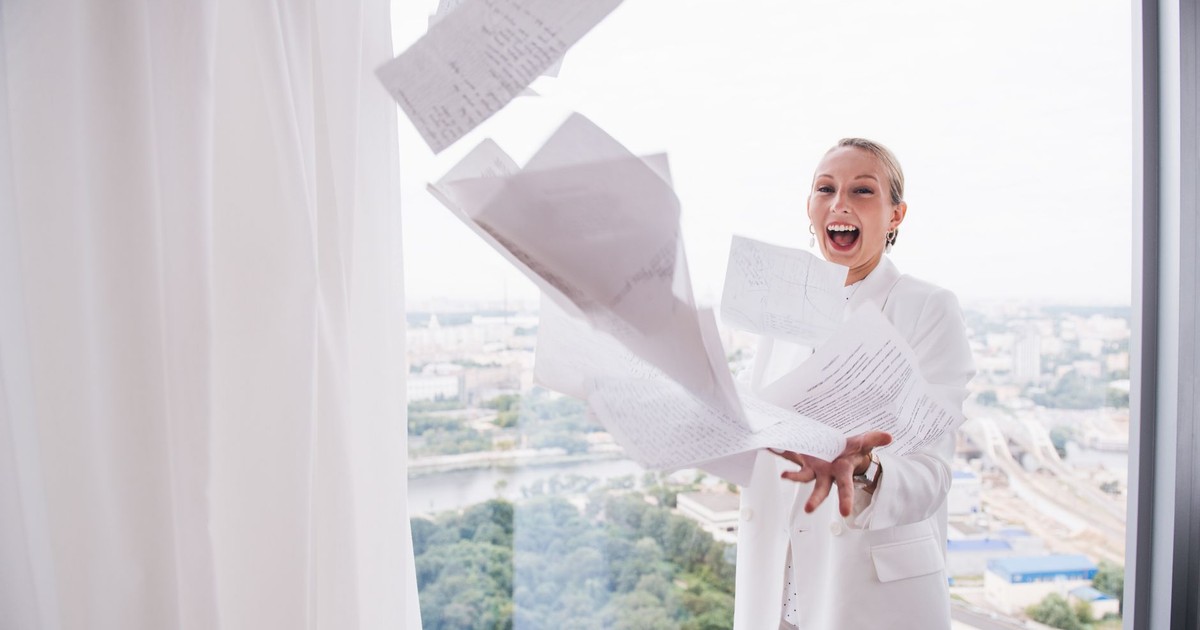 Businesswoman celebrating by tossing papers in a high-rise office with a city view.