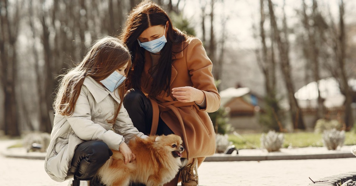 Cheerful daughter and mom in warm clothes and medical masks with little Pomeranian dog enjoying weekend in nature together