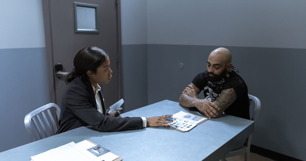 A woman interrogates a tattooed man in an interrogation room setting.