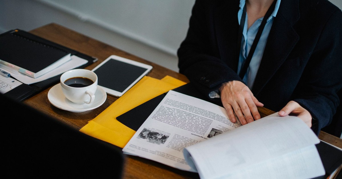 High angle of crop faceless businesswoman in formal clothes sitting at table with tablet and hot coffee and looking through documents