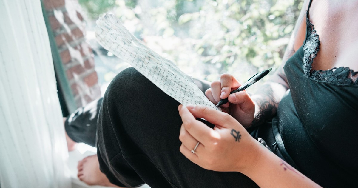 A woman sits on a windowsill writing on paper, bathed in sunlight.