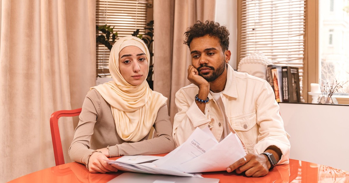 A couple in casual attire sits indoors at a table reviewing financial documents with a laptop.