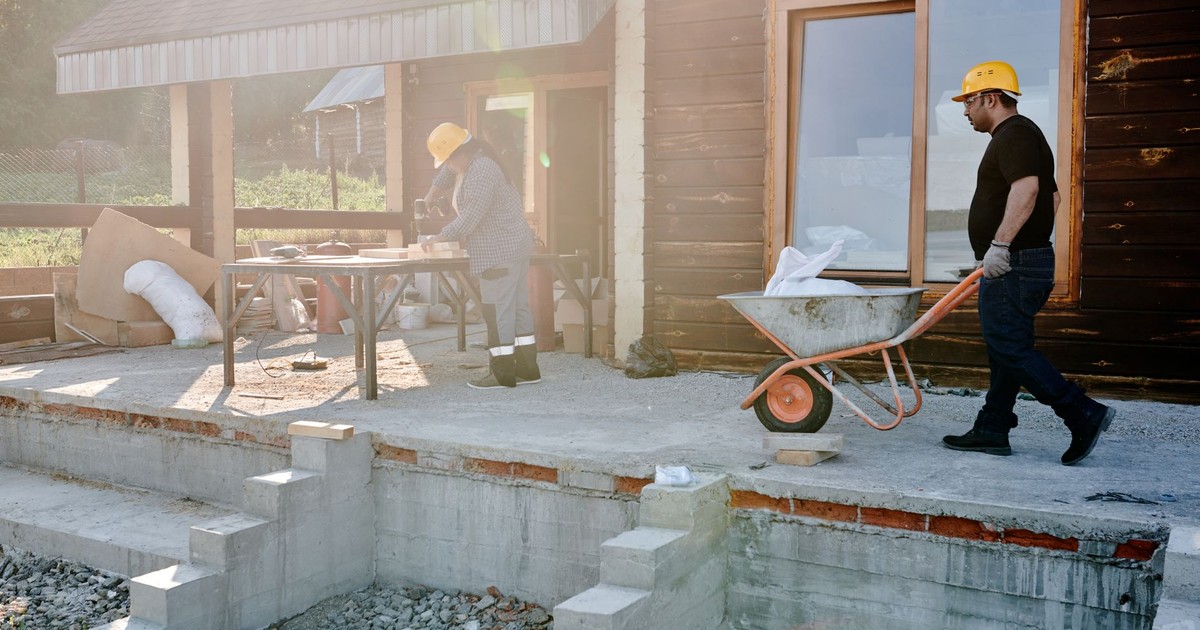 Two construction workers renovating a house exterior, carrying materials and working on woodwork.
