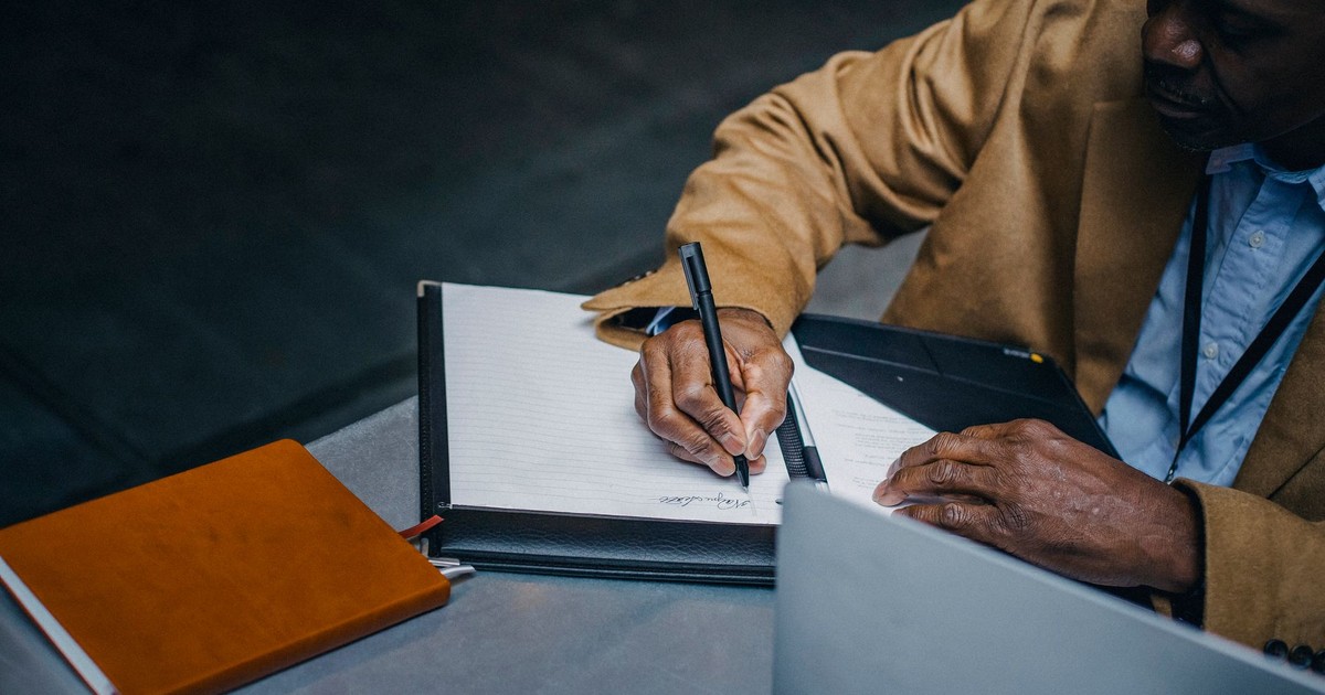 From above of crop black executive in suit writing on paper near laptop and diary while working at desk