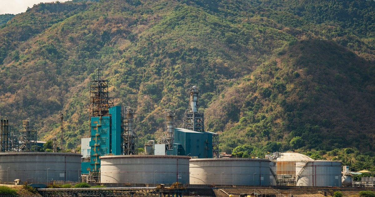 Industrial plant with large tanks and chimneys against a lush mountain backdrop.