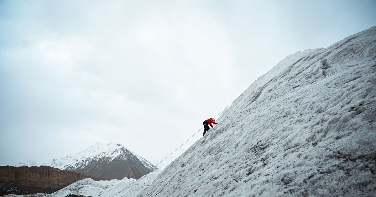 A climber in red jacket scaling a snowy mountain slope under a cloudy sky.