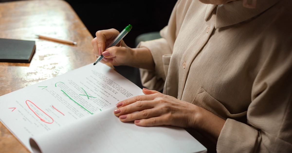 Close-up of a senior woman editing a manuscript at a desk with a green marker indoors.