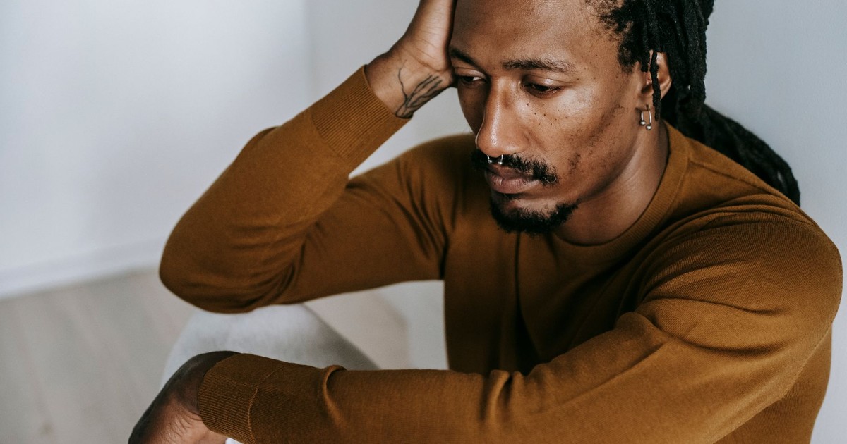 Crop young hopeless African American male with piercing touching head and looking down in house