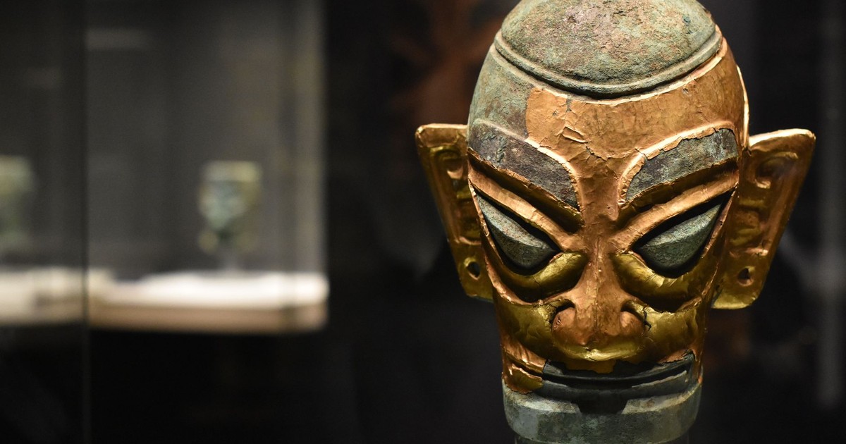 Close-up of an ancient bronze mask exhibited in a dimly lit museum display case.